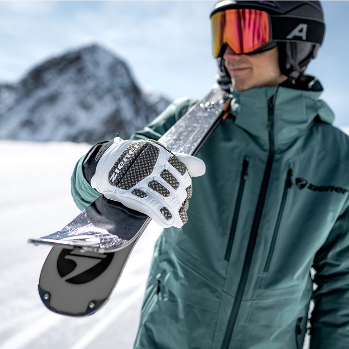 Skier in a green jacket carrying skis over the shoulder, with a white ZIENER racing glove in the foreground