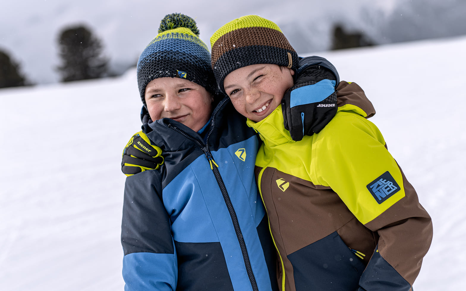 Zwei Jungen mit Mützen und bunten Skijacken umarmen sich im Schnee