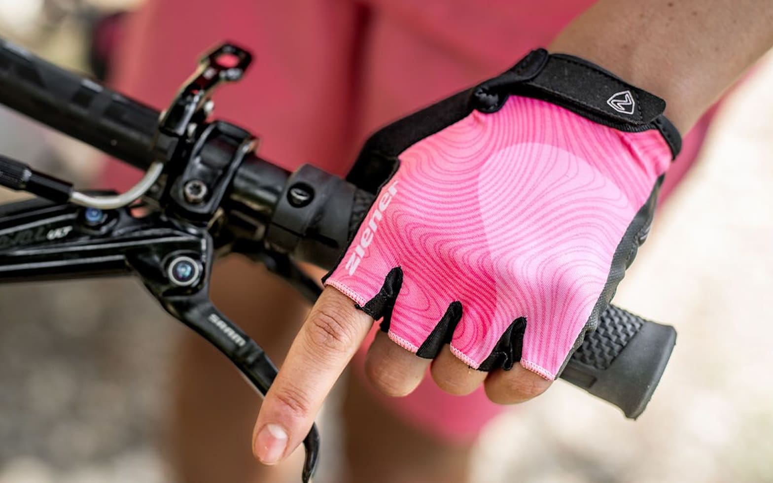 Nahaufnahme einer Hand im pinken ZIENER Kurzfinger-Bikehandschuh am Lenker